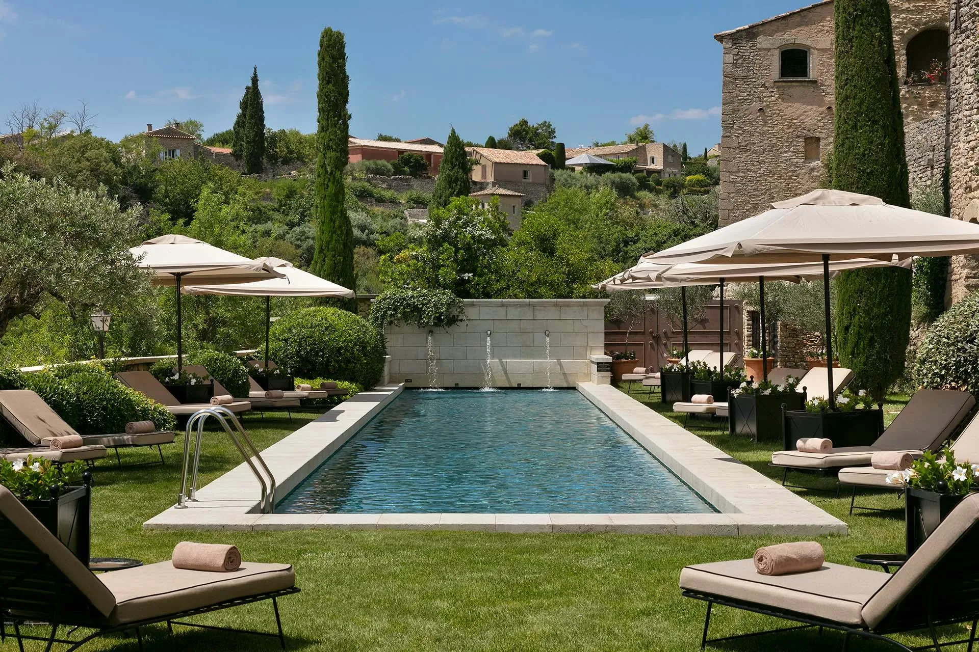 Rectangular pool lined with loungers and white umbrellas surrounded by stone walls and cypress