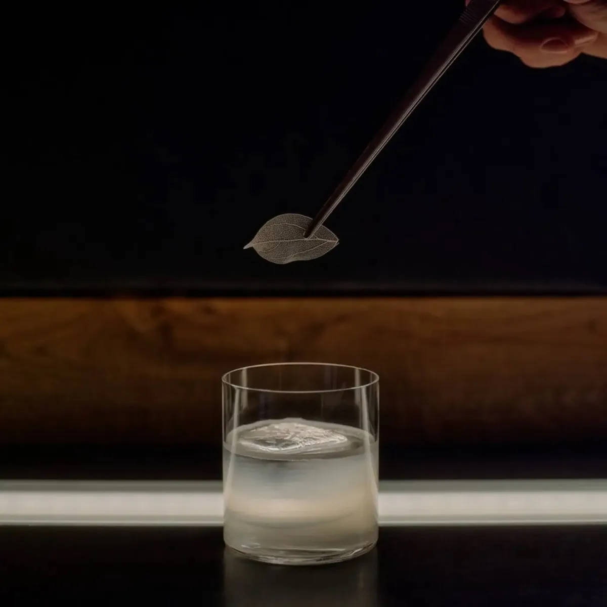 Cocktail garnished with a dried leaf, poured tableside over ice in a low rocks glass