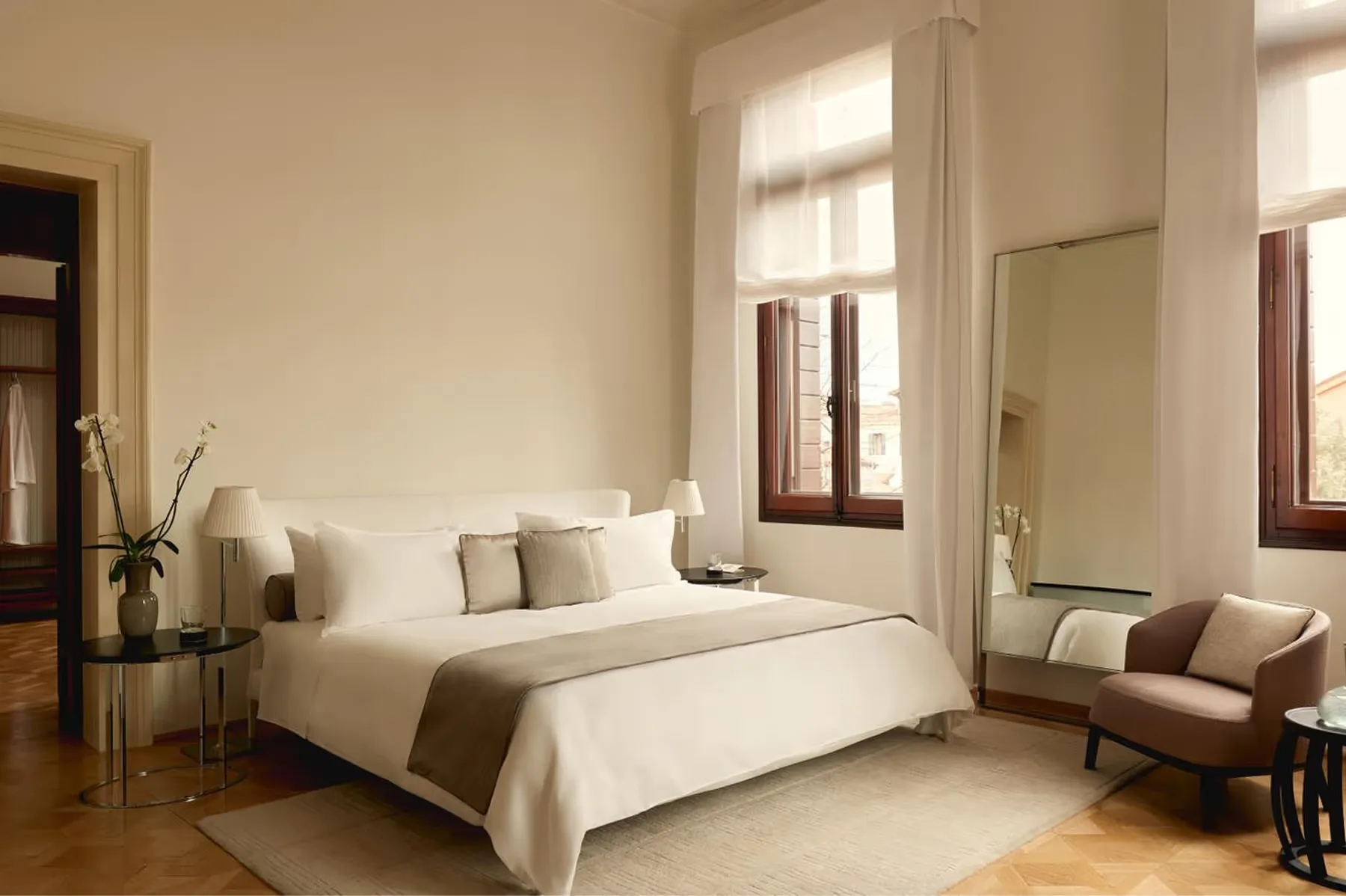 A bright bedroom with neutral linens, brown leather seating, tall windows with natural light, and wooden flooring