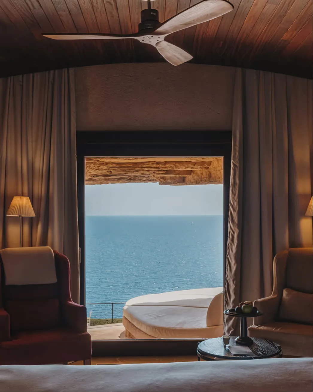 A modern ceiling fan rotates above cream curtains, a white soaking tub, and a seascape beyond