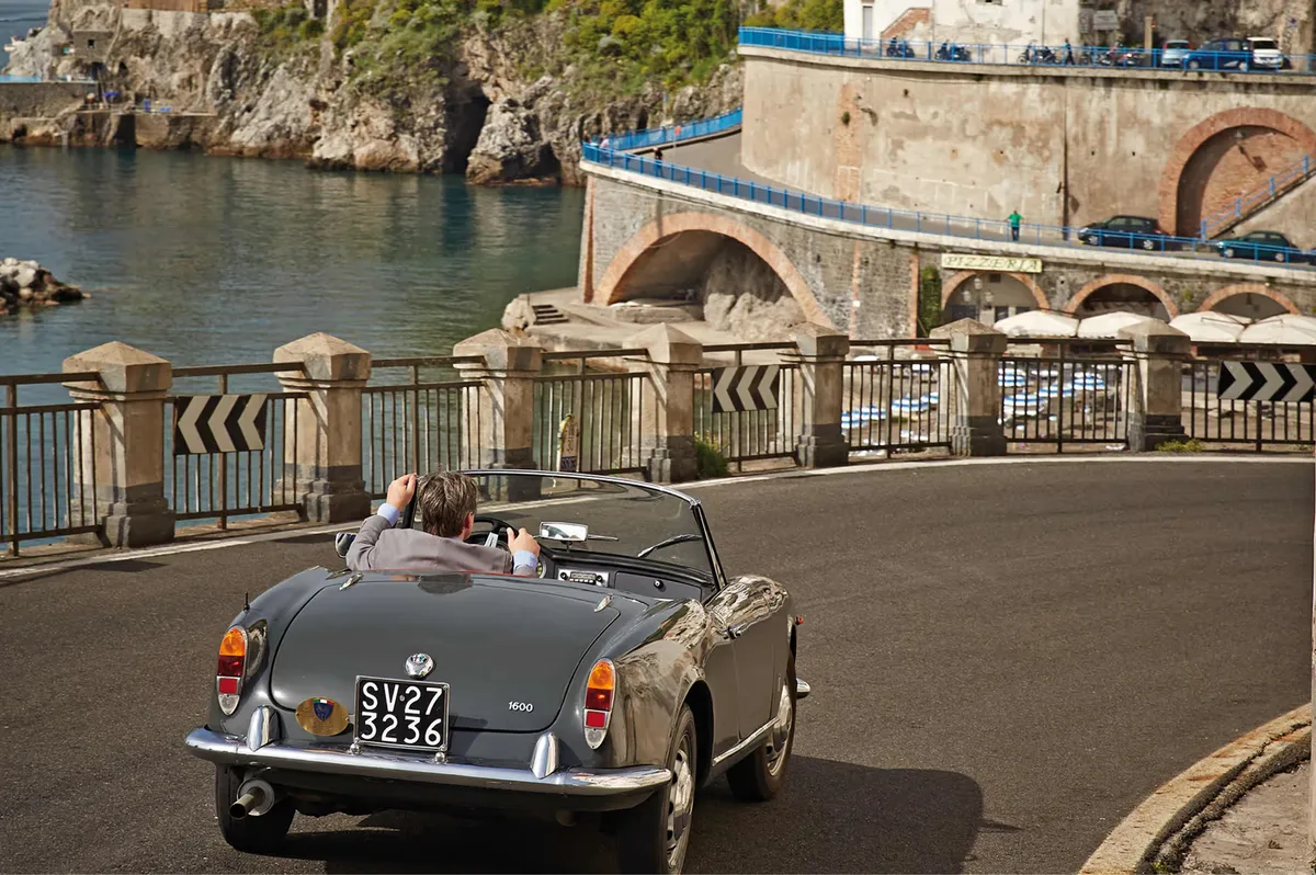 Vintage convertible car parked on coastal bridge with waterfront buildings and boats