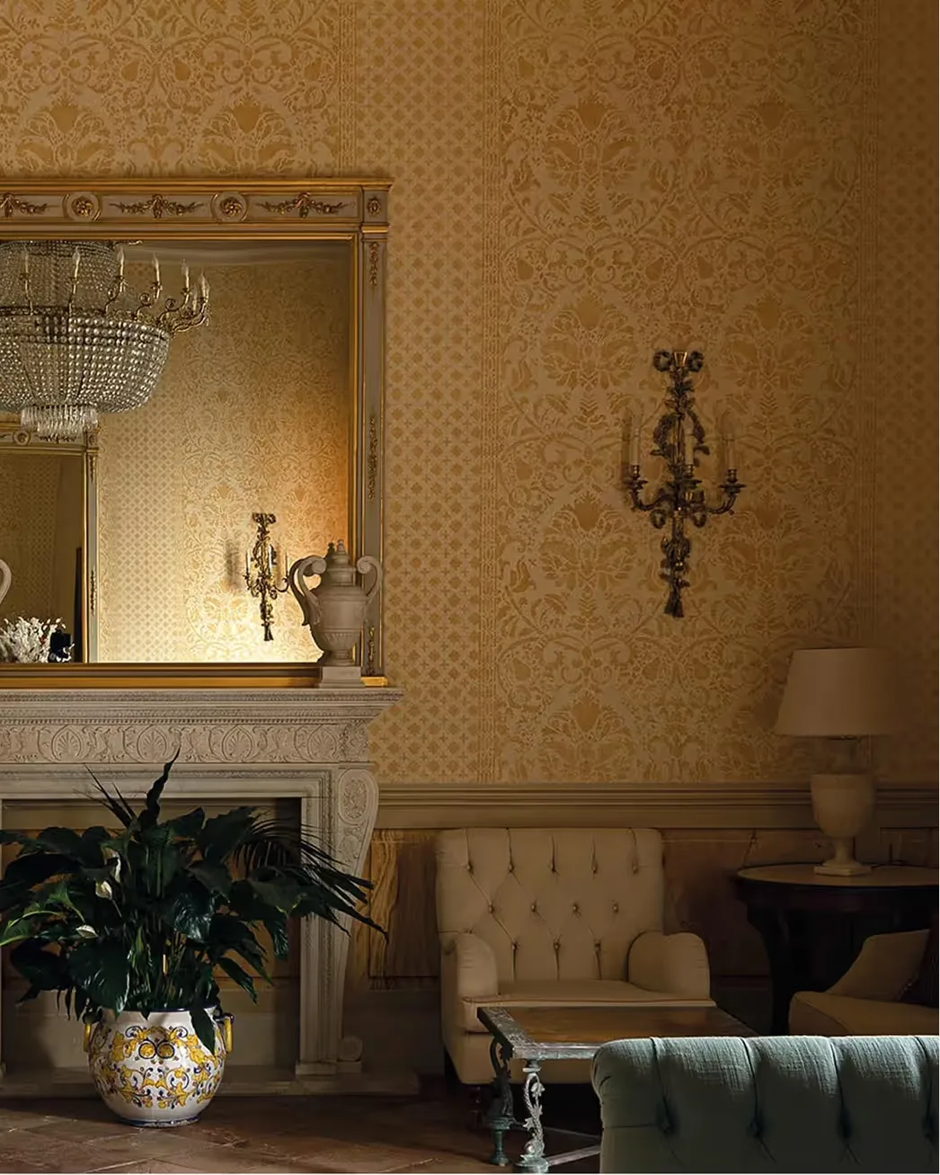 Golden patterned wall with ornate mirror, fireplace, chandelier, cream upholstered chair, and potted plant