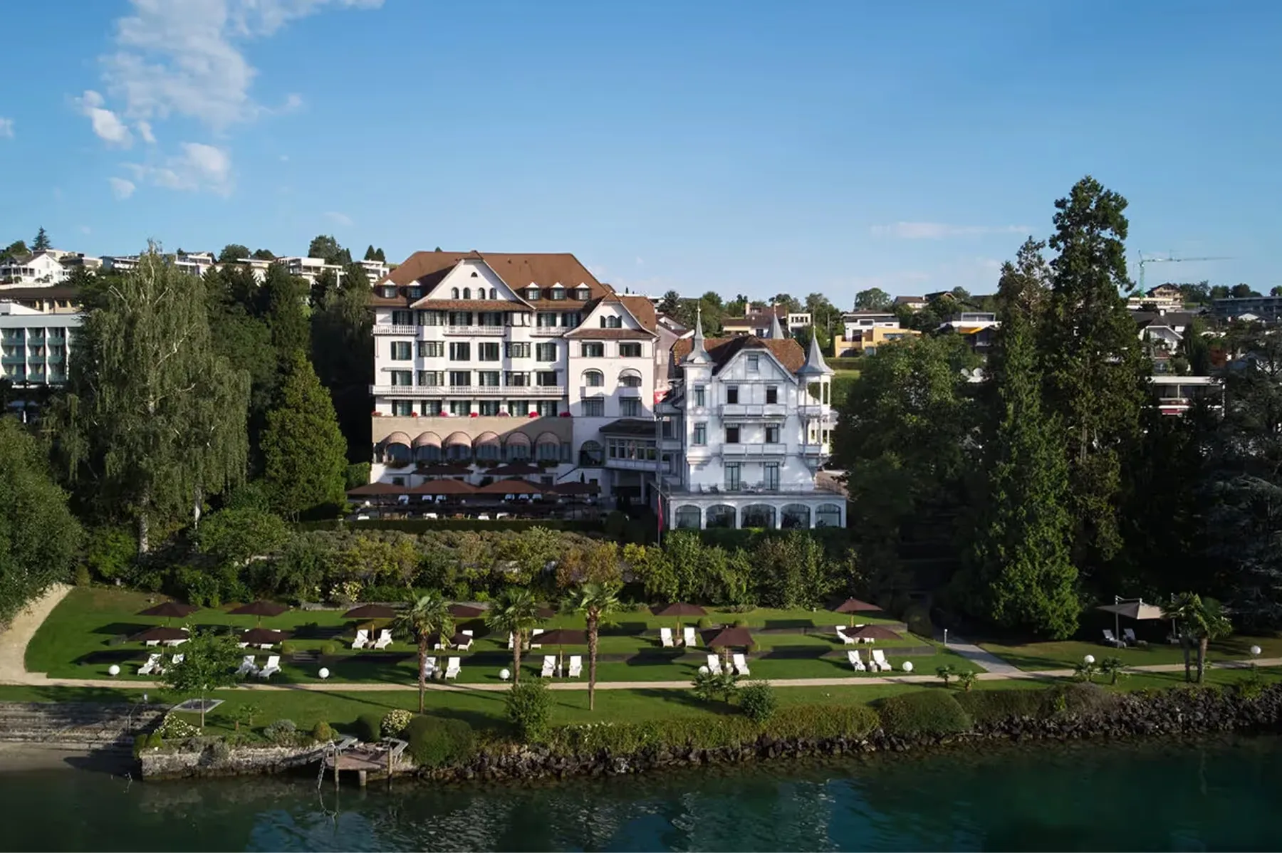 Pale stone palace with pitched roofs, turret tower, and cypress trees overlooking Lake Lucerne and manicured gardens