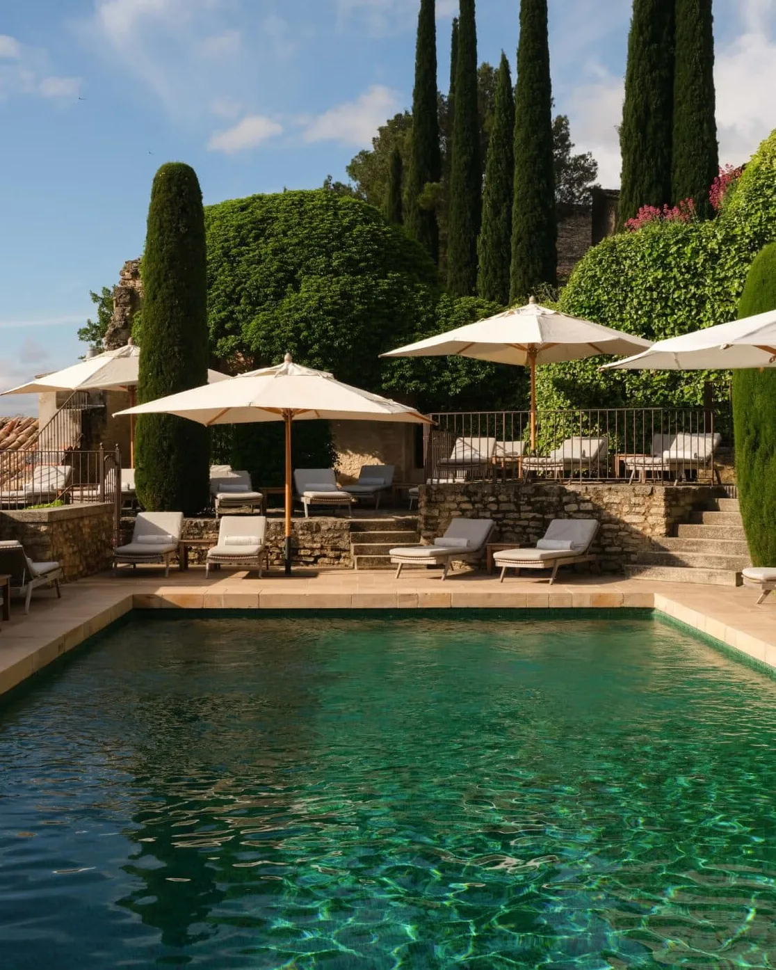A terrace with sun umbrellas, lounge chairs, and a turquoise pool set against cypress trees