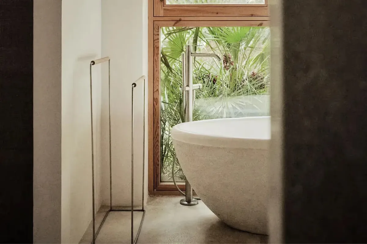 Freestanding sculptural bathtub positioned beside metal-frame window overlooking verdant landscape