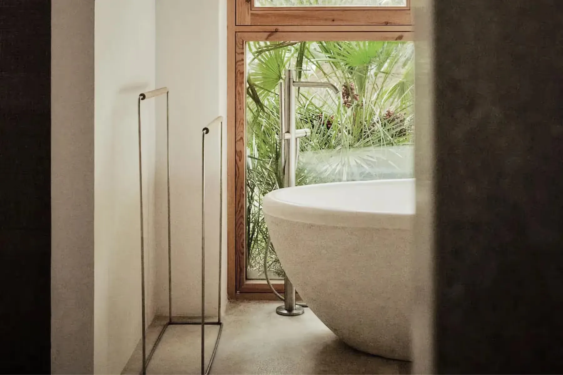 Freestanding sculptural bathtub positioned beside metal-frame window overlooking verdant landscape