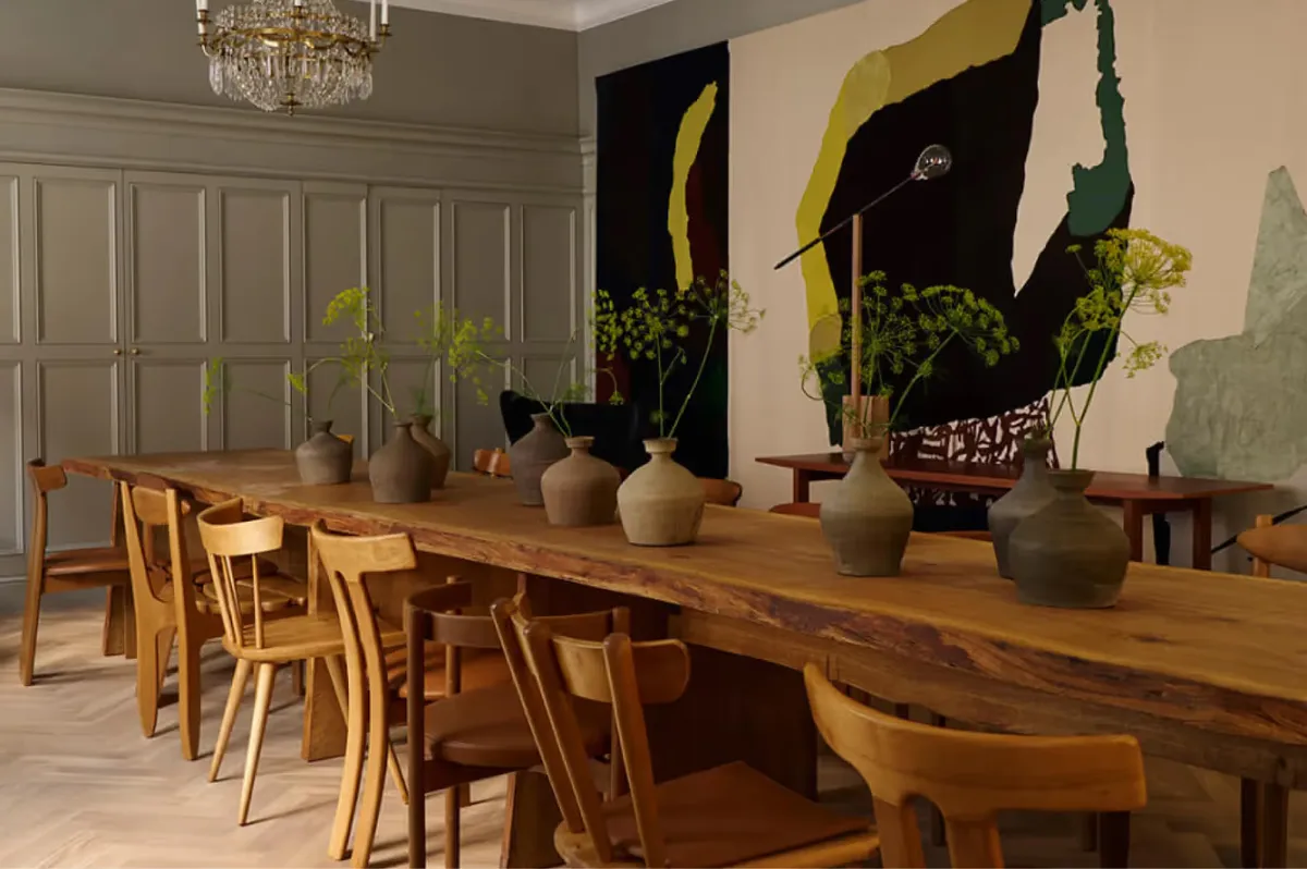 A dining room with wooden table and chairs, potted plants, and dark artwork on neutral walls