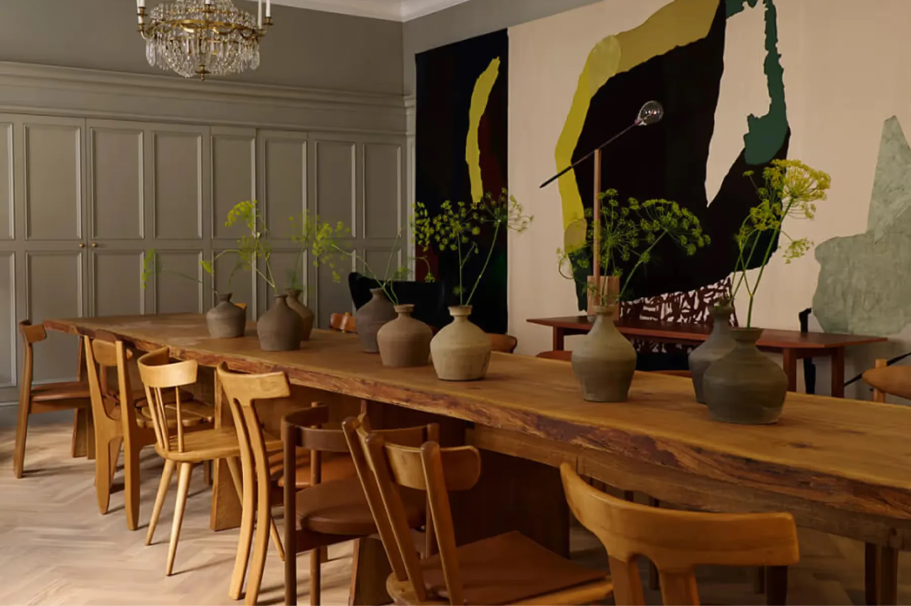 A dining room with wooden table and chairs, potted plants, and dark artwork on neutral walls
