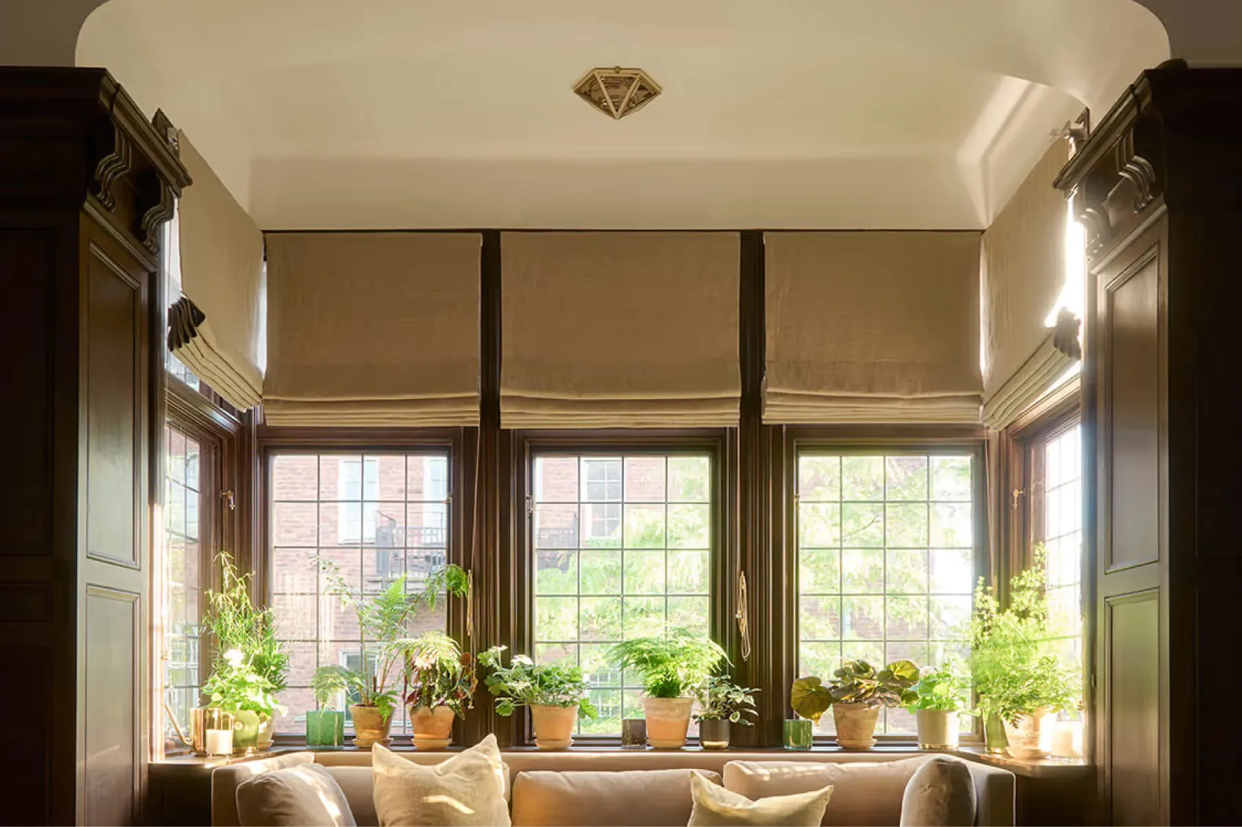 A window seat with large arched windows, cushions, and potted plants overlooking the exterior