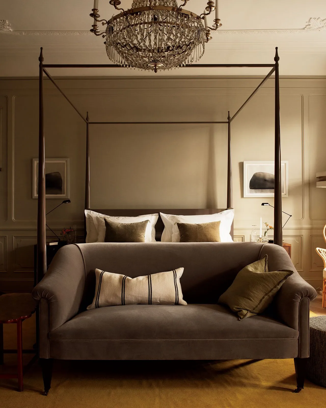 A canopy bed with four dark posts, cream bedding, and ornate crystal chandelier overhead