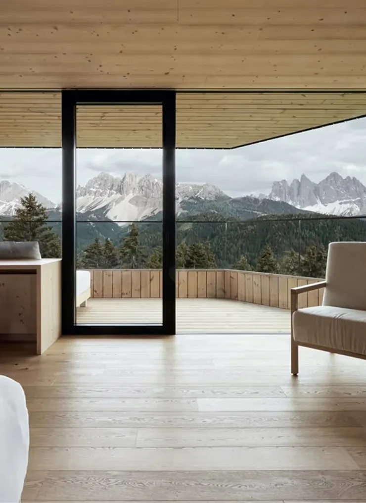 Suite with large black-framed window opening to a wooden deck overlooking forested mountains under pale sky