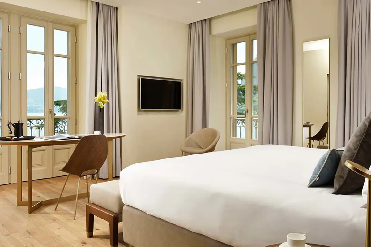 A spacious bedroom with light wood floors, large windows, and a desk