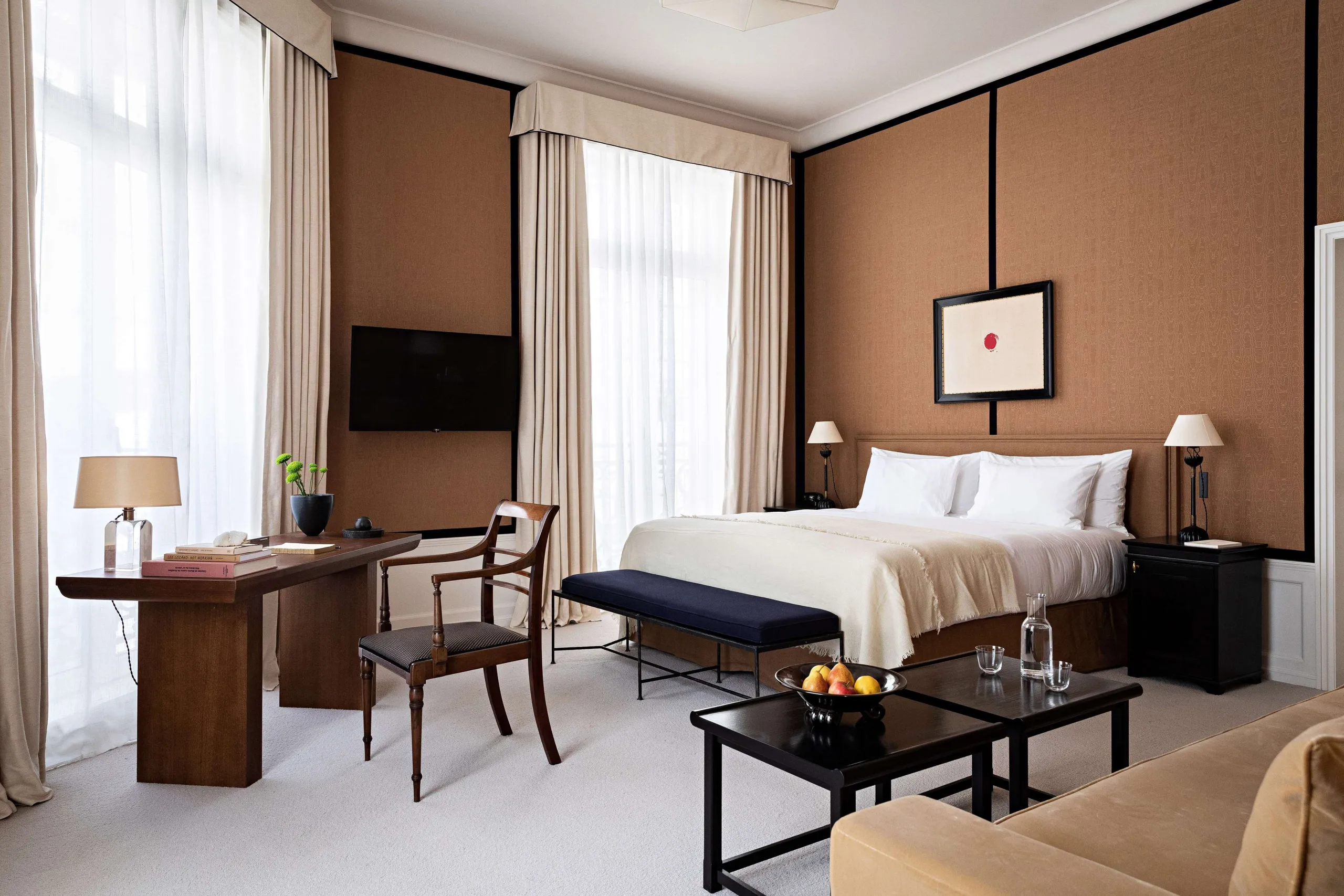 A bedroom with tan and brown tones, wood furnishings, and tall windows