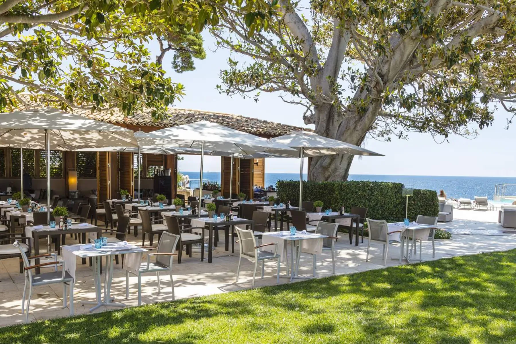 Dining tables arranged under shaded structures with Mediterranean seascape