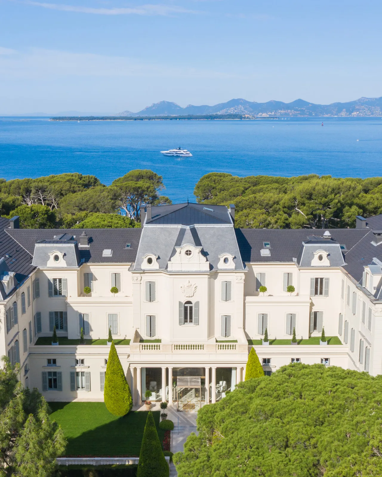 The classical facade of a cream-colored resort hotel overlooking a turquoise cove, with the Mediterranean Sea and distant coastline