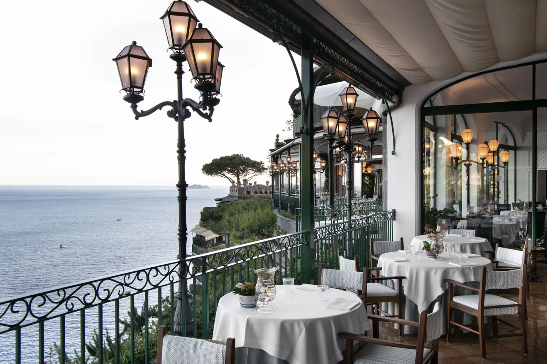 Elegant covered patio dining with white tablecloths, brass lanterns, railing, and sea views