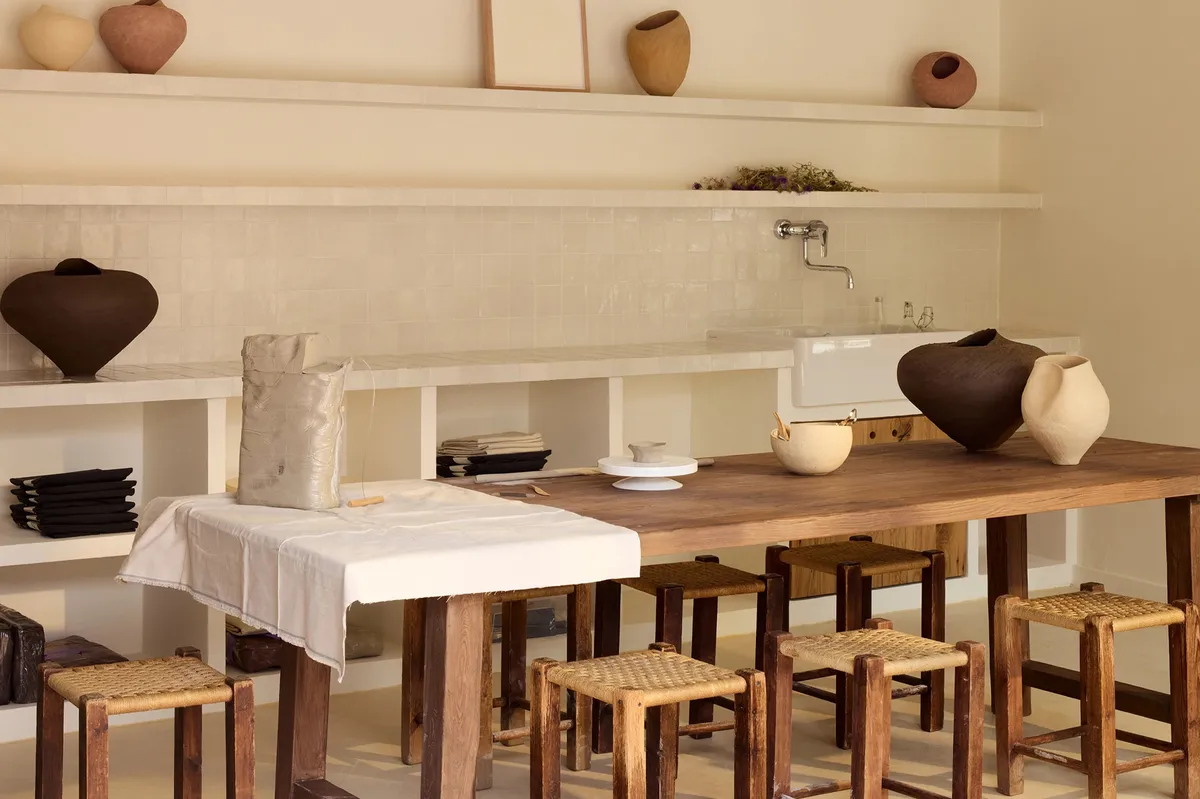 Dark and pale ceramic vessels line a wooden table beneath a tiled backsplash and shelf