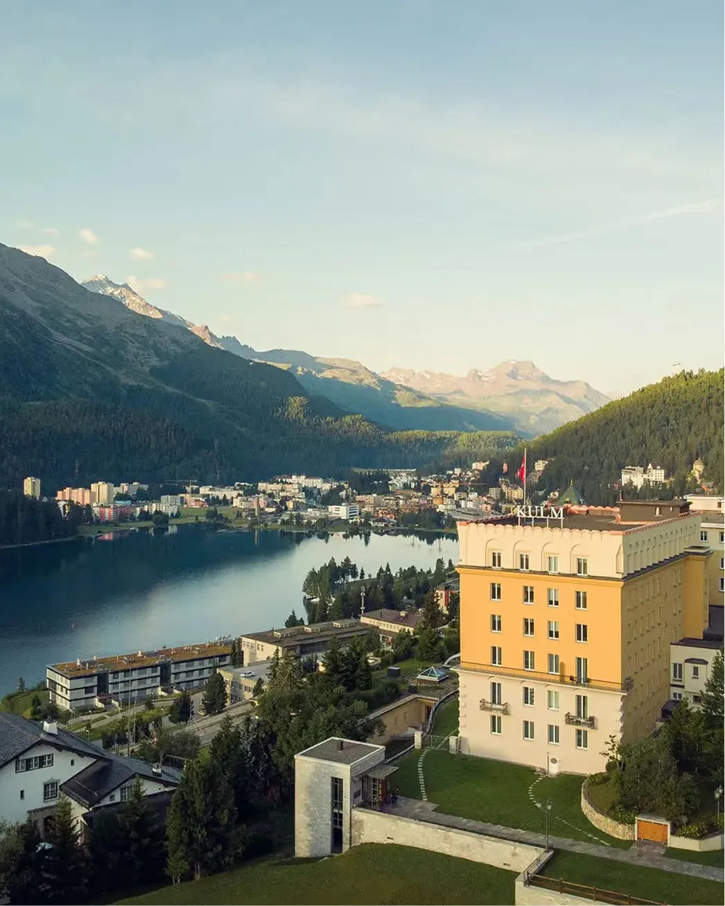 The hotel’s yellow building towers above the lakeshore with snow-capped mountains