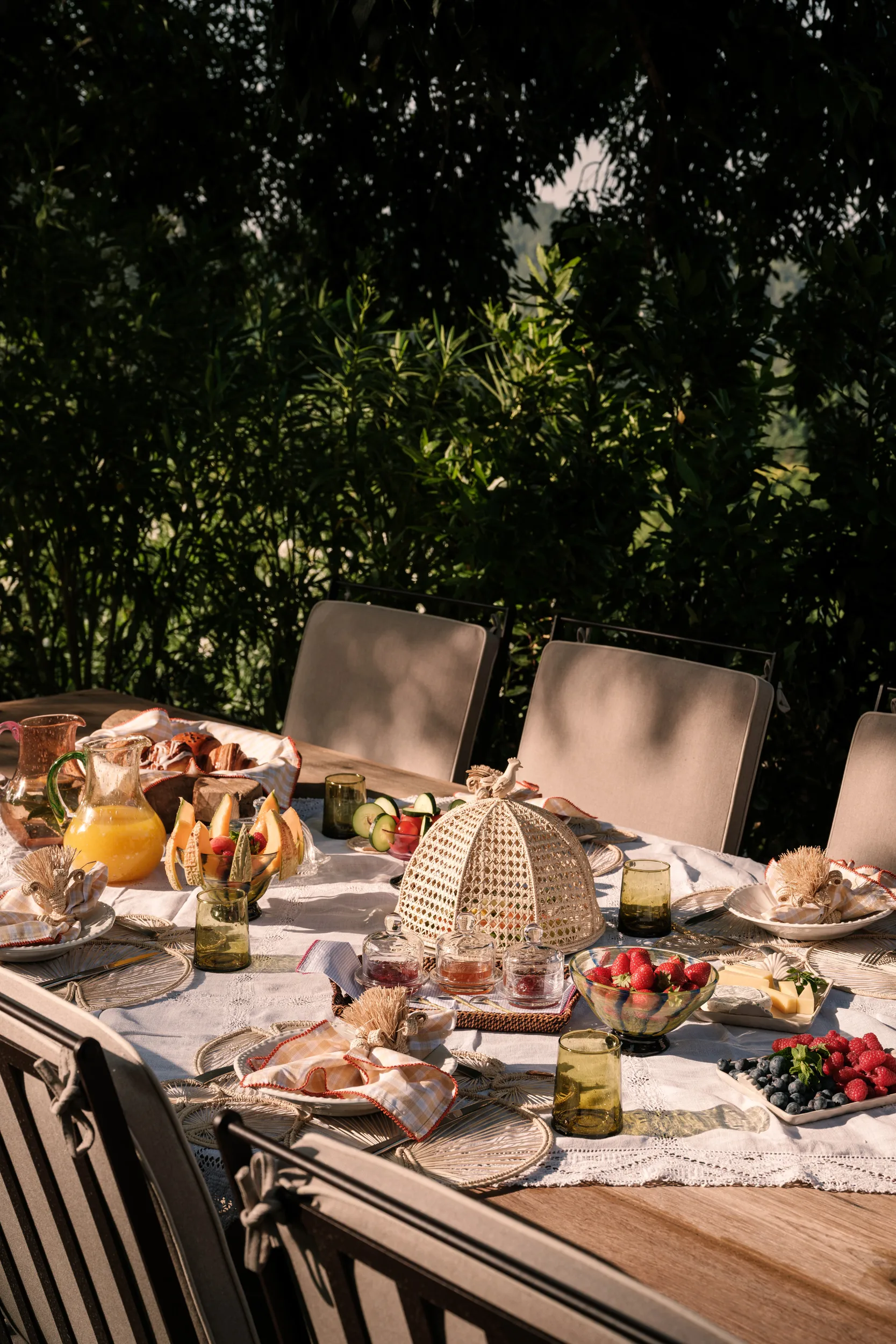 Outdoor breakfast table set with pastries and fruit beneath woven shade sail, lush green foliage, dappled light