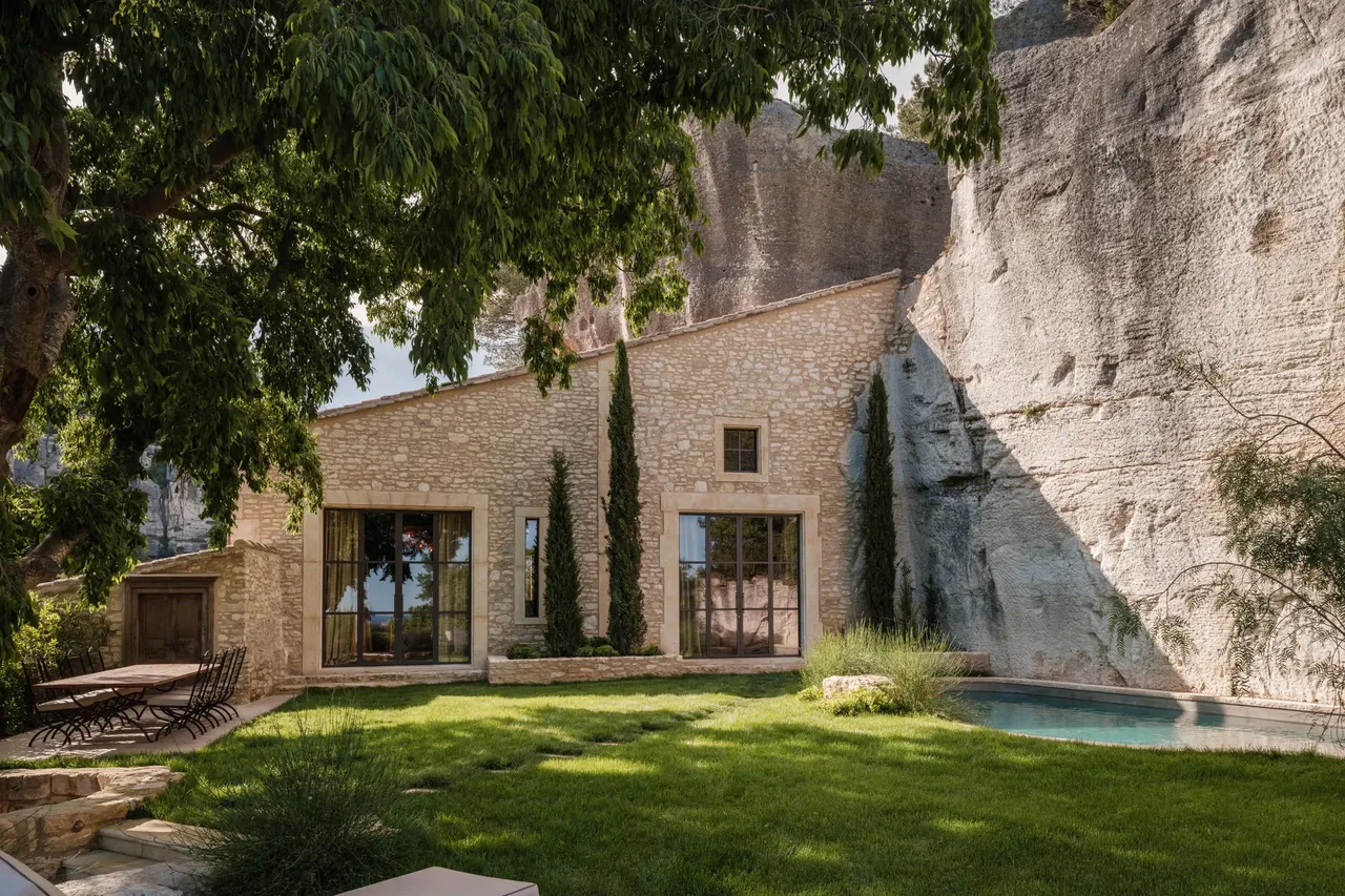 Stone villa with modern glass doors, wooden pergola, grass courtyard, pale turquoise pool, limestone cliff backdrop
