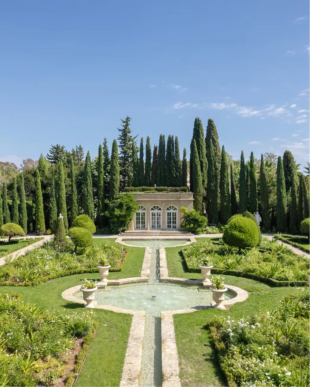 Pale stone château with arched loggia, cypress trees, and manicured gardens against clear sky