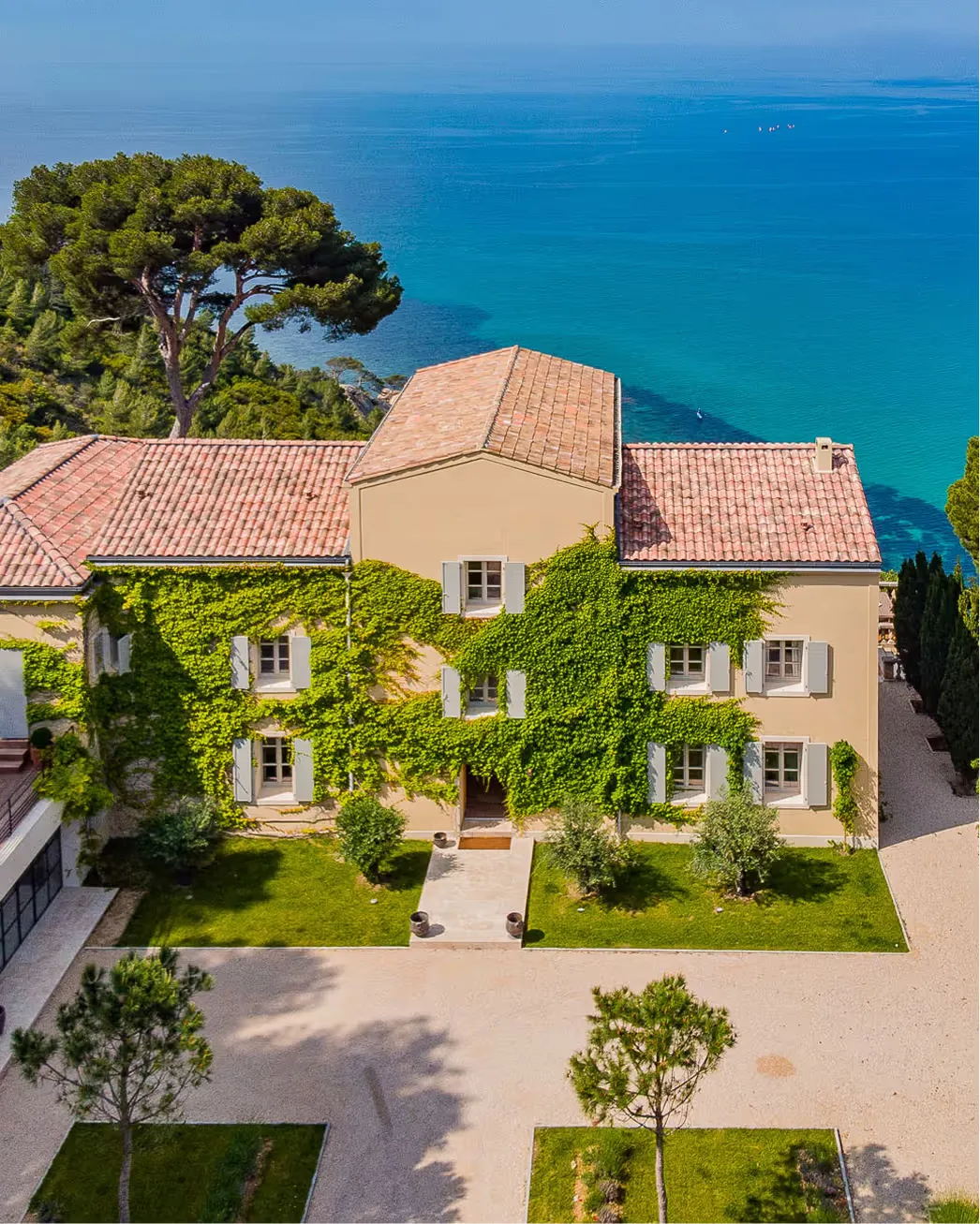 Ochre-colored manor with ivy-covered walls and shutters, sea beyond, set in Mediterranean landscape