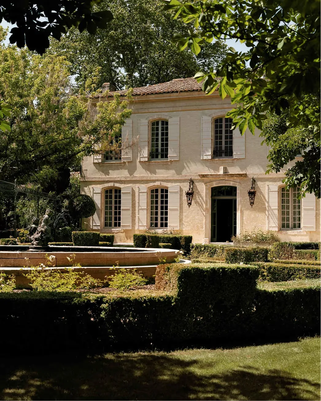 Pale stone manor framed by dappled olive tree foliage and hedged borders