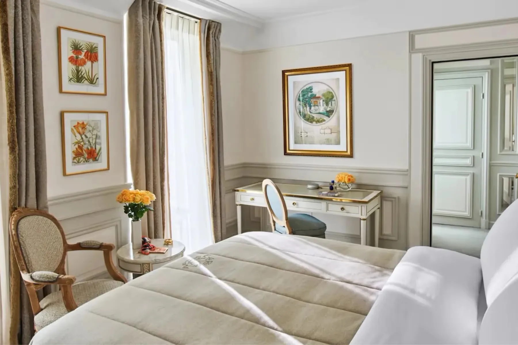 A suite with queen bed, pale furnishings, framed prints, and tall windows with views