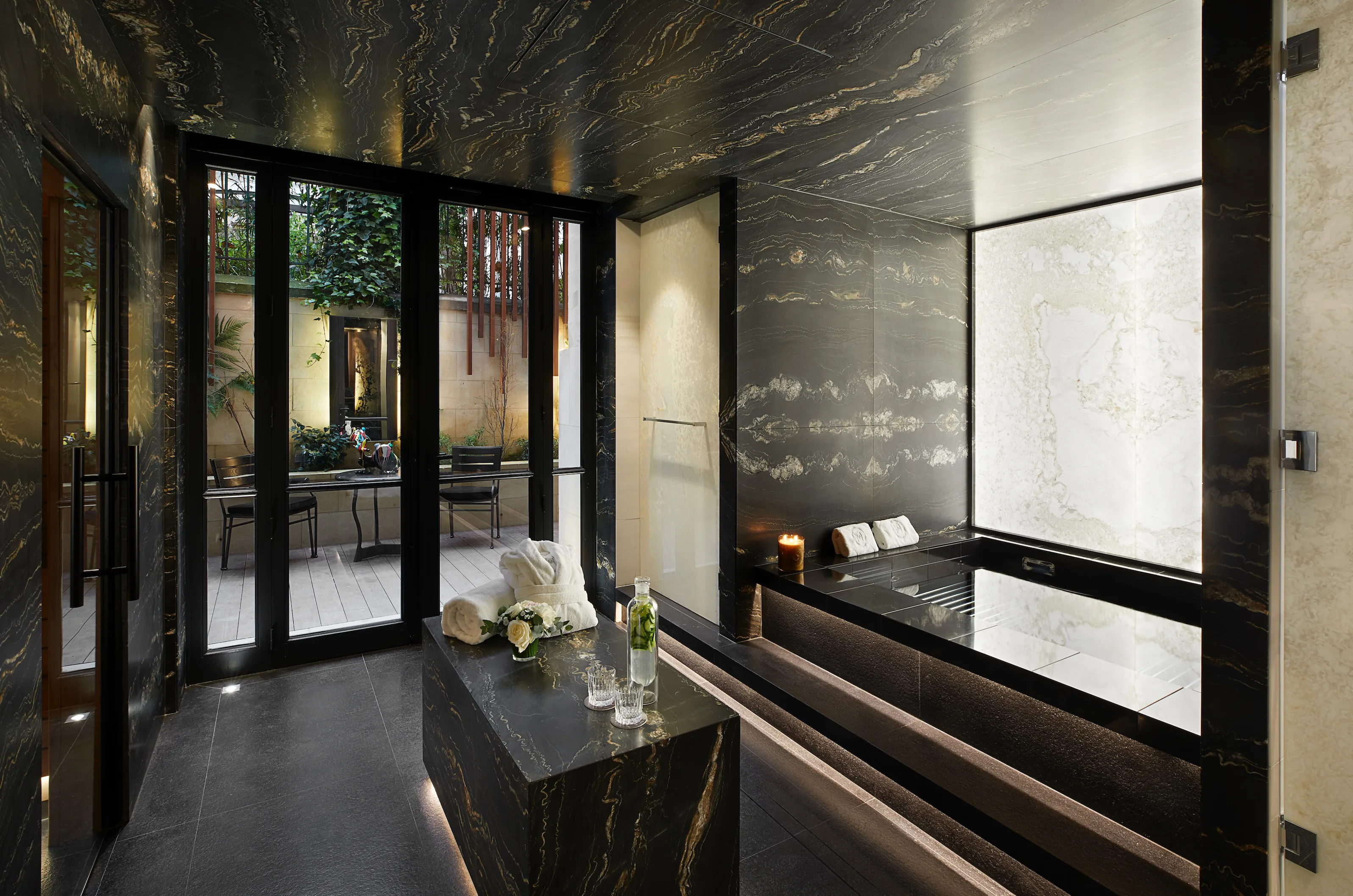 A dark spa interior with glass doors opening to a garden, pale marble walls, and modern furnishings