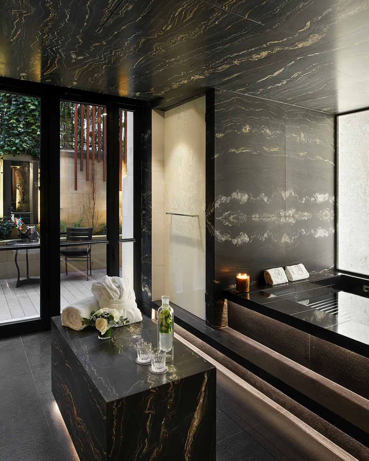 A spa interior with a dramatic black marble wall, glass doors, light-colored furnishings, and patterned ceiling