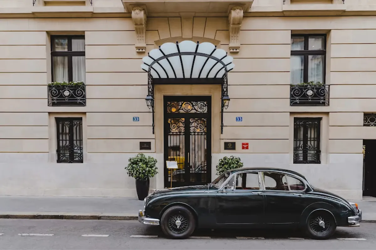 A classical beige facade with arched entry, black-trimmed windows, and a black vintage car parked outside