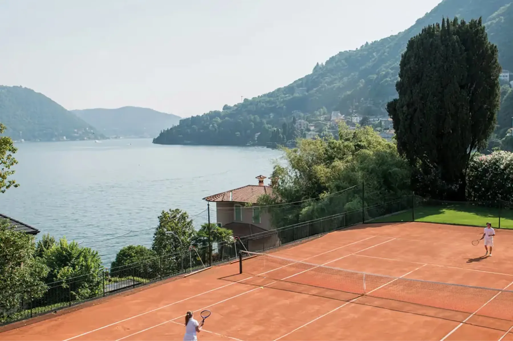 A clay tennis court with lake and mountain scenery on the opposite shore