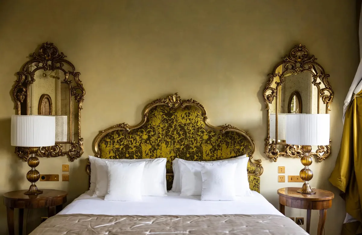 A bed with ornate gold headboard flanked by mirrored bedside tables and sconces