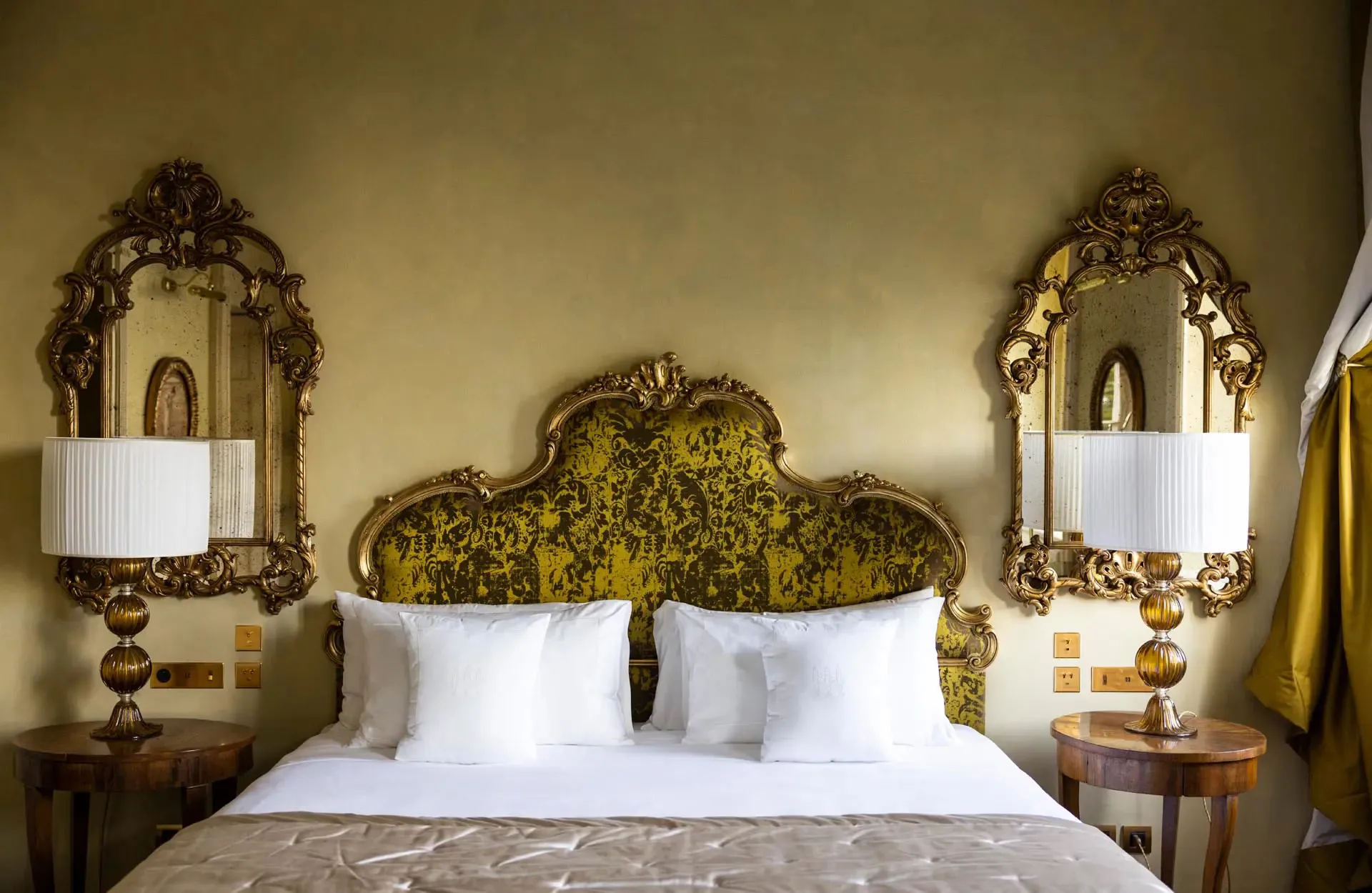 A bed with ornate gold headboard flanked by mirrored bedside tables and sconces