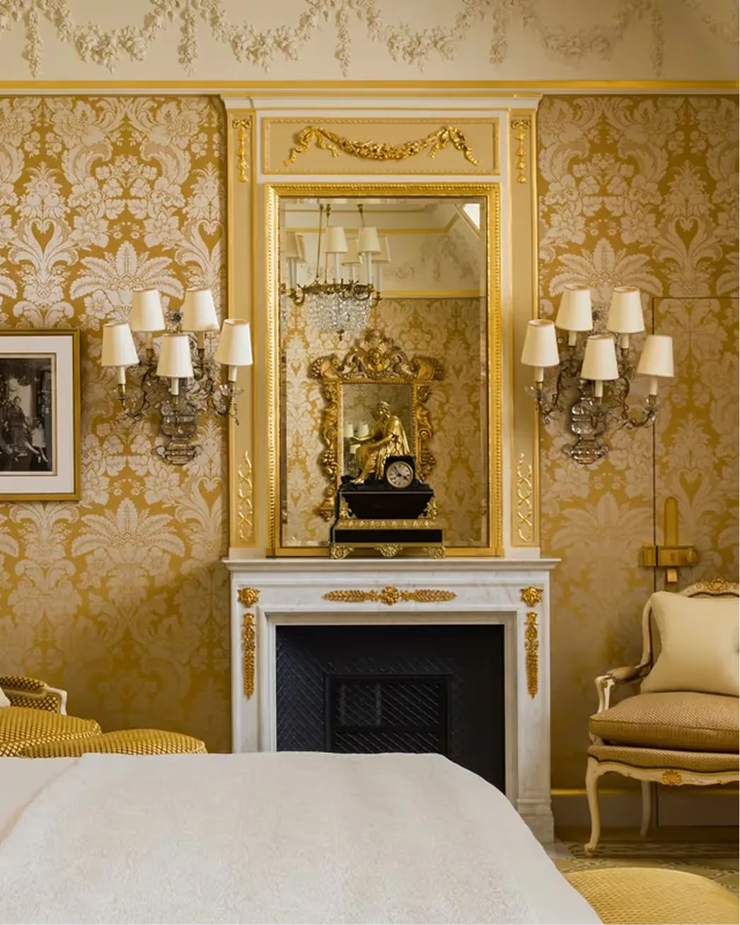 Gilt-framed mirror above a marble fireplace with candelabras against damask wallpaper