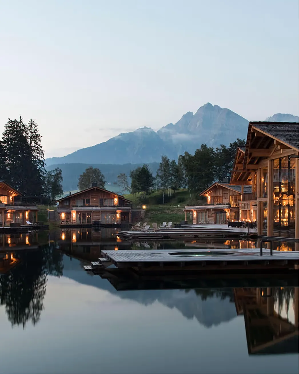 The property lit at dusk beside a reflective lake with mountains behind