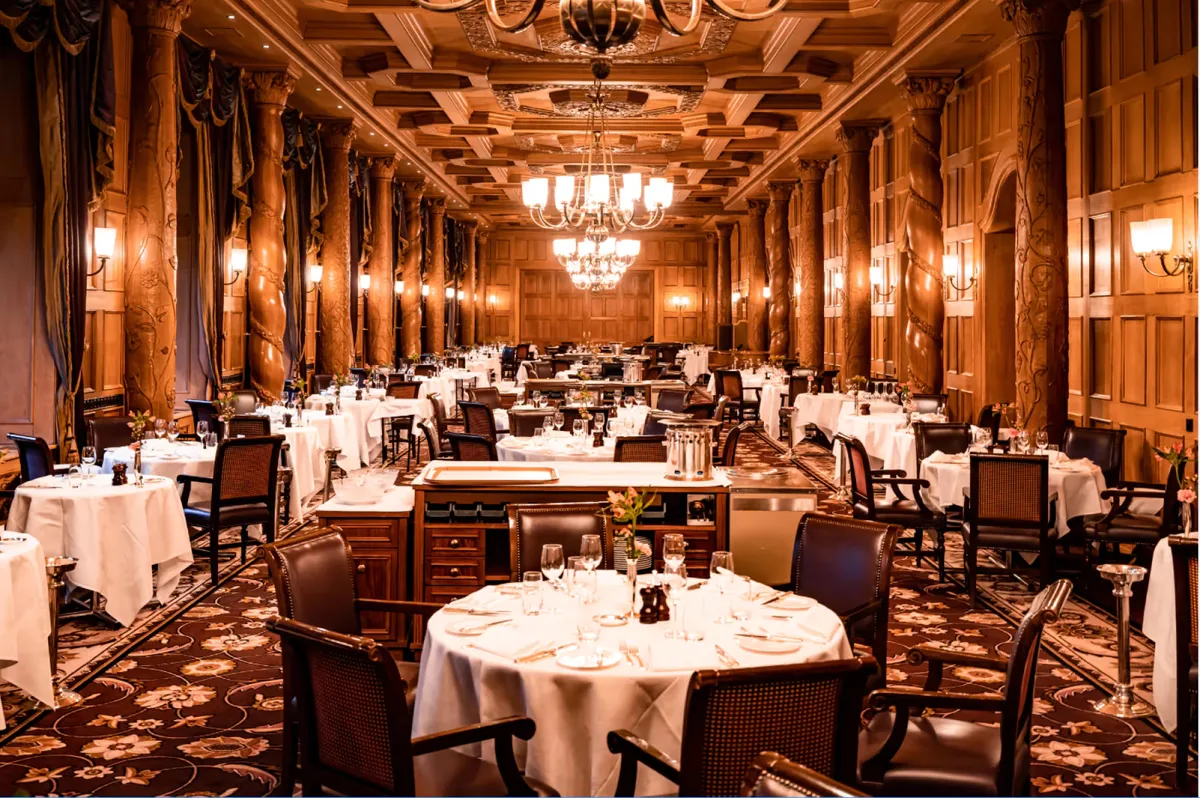 A grand dining hall with ornate golden ceiling, crystal chandeliers, and formally set white-draped tables