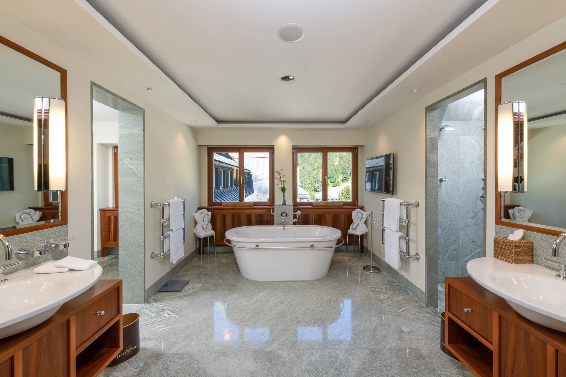 A spa bathroom with dual sinks, a soaking tub, large windows, and neutral stone finishes