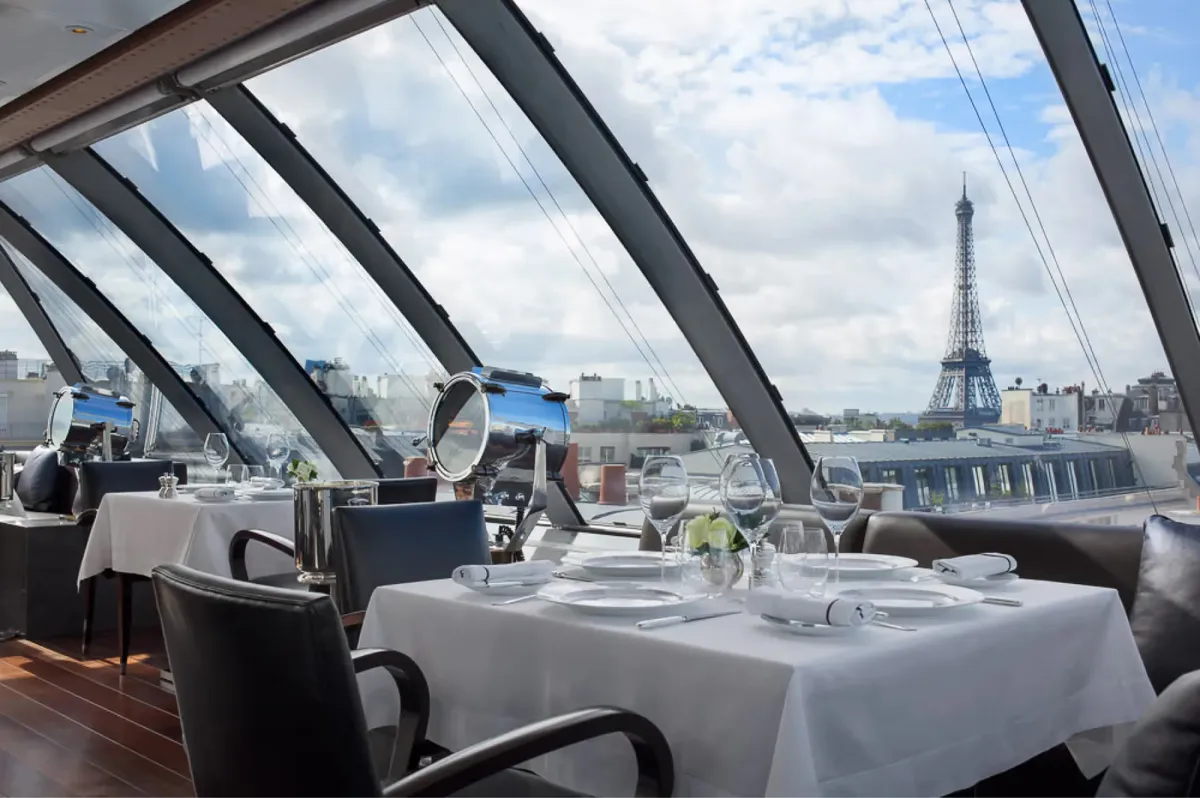 Rooftop dining terrace with glass panels, white tablecloths, and panoramic views of the Eiffel Tower