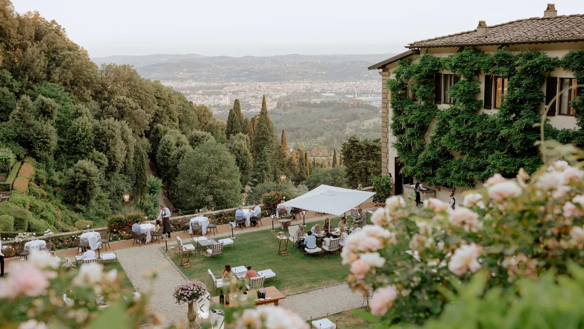 An afternoon garden party with guests at scattered tables, white umbrellas, and valley views beyond