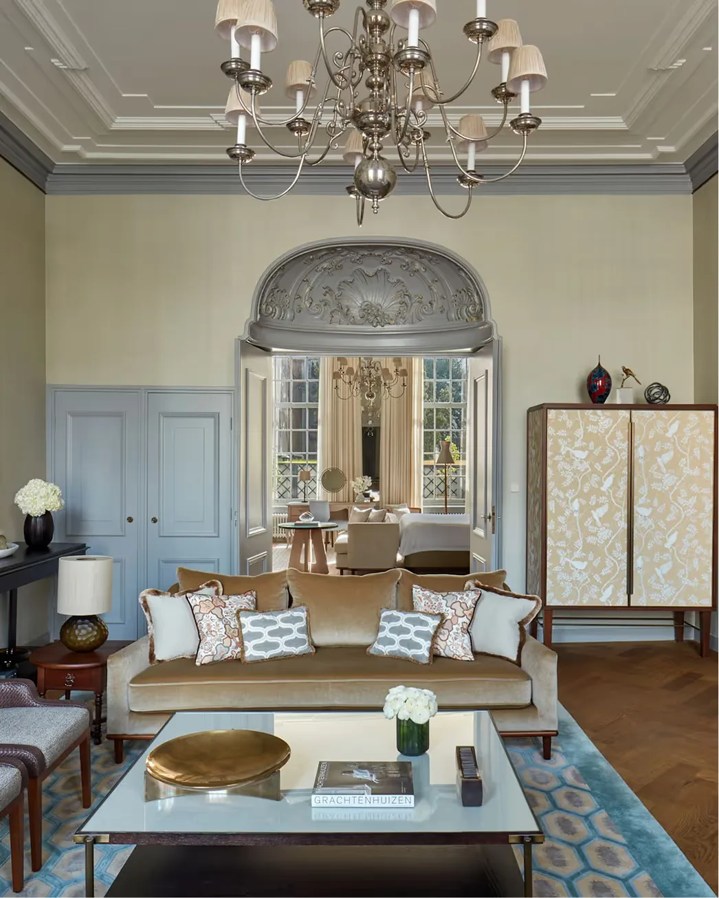 A blue-shuttered room with a crystal chandelier, sofa with patterned cushions, and tall windows framing a canal view